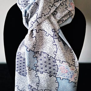 May include: A white scarf with a repeating pattern of blue and pink floral and geometric shapes. The scarf is draped over a black form, showcasing its design and texture. The scarf's design includes a mix of light blue, dark blue, and pink elements.