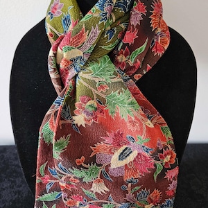 May include: A textured scarf featuring a vibrant floral pattern with deep red, olive green, blue, and purple hues. The fabric displays a dense, intricate botanical design with detailed petals and leaves covering the entire surface of the accessory.