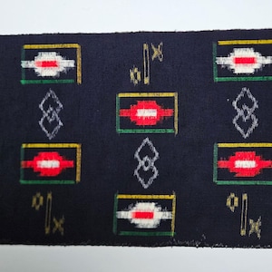 May include: Rolled navy blue fabric with a repeating pattern of red, white, and green rectangles outlined in yellow. The design includes white diamond shapes and gold symbols. The fabric is likely used for upholstery or crafts.