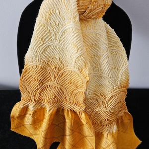 May include: A yellow and cream-colored scarf displayed on a black mannequin. The scarf features a textured, patterned design with a ruffled edge. The fabric transitions from cream to yellow, with a geometric pattern on the bottom.