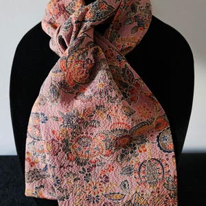 May include: A pink scarf with an intricate floral pattern in shades of red, orange, blue, and brown. The scarf is draped over a black form, showcasing its detailed design and textured fabric. This accessory adds a touch of elegance.