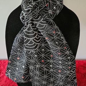 May include: Black scarf with a white geometric pattern and small orange accents. The scarf is draped over a black mannequin. The pattern includes circles, diamonds, and lines, creating a detailed design. The scarf is made of a soft material.