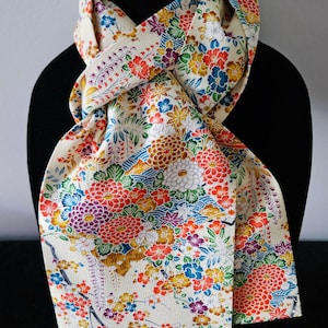 May include: Cream-colored scarf with a vibrant floral pattern in red, blue, purple, and yellow. The scarf is draped over a black mannequin, showcasing the intricate design and color palette. The fabric appears to be silk or a similar lightweight material.
