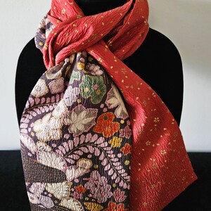 May include: A two-sided scarf displayed on a black mannequin. One side is a textured red with small gold accents, the other features a floral pattern in shades of purple, pink, orange, and green. The scarf is made of a soft, flowing fabric.