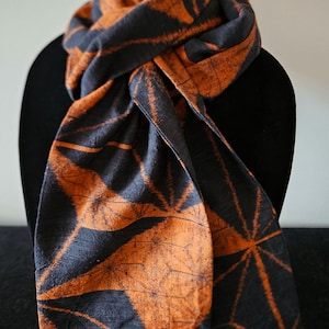 May include: A black and orange scarf with a geometric leaf pattern. The scarf is draped over a black form, showcasing the intricate design and color contrast. The fabric appears soft and textured.