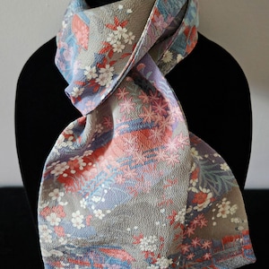 May include: A textured scarf featuring a grey base with a repeating pattern of white cherry blossoms, pink maple leaves, and blue geometric shapes. The fabric has a crinkled, embossed surface texture throughout, with a soft drape and folded design.