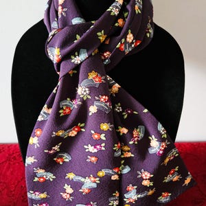 May include: A deep purple scarf featuring a repeating pattern of small, colorful flowers and grey hair combs. The fabric has a textured surface and is draped in a knot, displaying the floral and comb motifs across the entire length of the material.