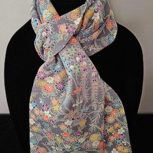 May include: A grey scarf featuring a dense, colorful floral pattern with small blossoms in shades of orange, yellow, blue, purple, and white. The fabric has a textured surface and is draped in a loop, displaying a repeating botanical design.