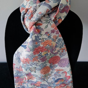 May include: A cream-colored scarf featuring a textured surface and a dense, colorful floral pattern. The design includes various flowers in shades of red, orange, purple, and blue, interspersed with green leaves and stems across the fabric.