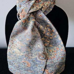 May include: A patterned scarf with a floral design in shades of blue, beige, and rust. The scarf is draped over a black mannequin, showcasing its intricate details and color palette. The fabric appears to have a textured surface.