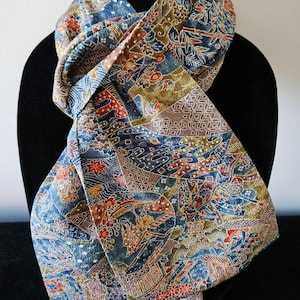 May include: A patterned scarf with a complex design in shades of blue, brown, red, and gold. The scarf is draped over a black form, showcasing its intricate details and textures. The fabric appears to be soft and flowing, suitable for wear.