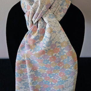 May include: A floral patterned scarf in pastel colors of pink, blue, yellow, and purple. The scarf is draped over a black mannequin, showcasing its design. The fabric has a delicate, textured appearance, with a repeating floral pattern.