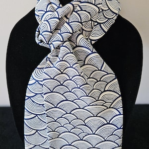 May include: A white and navy blue scarf with a repeating wave pattern. The scarf is draped over a black mannequin, showcasing the design. The pattern is a series of curved lines, creating a textured appearance. The scarf is made of a soft, flowing fabric.