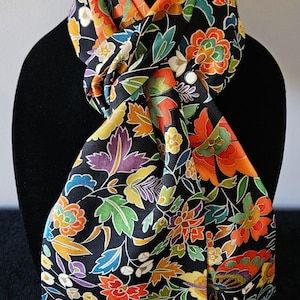 May include: A black scarf with a vibrant floral pattern in shades of orange, yellow, green, purple, and blue. The scarf is draped over a black mannequin, showcasing its design and texture. The scarf's intricate details are visible.