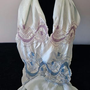 May include: A pale mint green scarf featuring a subtle tonal wave pattern throughout. The fabric is gathered with intricate embroidery in shades of purple and blue, creating a textured, ruched effect across the center and upper sections.