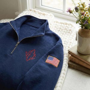 May include: Navy blue quarter-zip sweatshirt with a red embroidered logo and an American flag patch on the sleeve. The sweatshirt has a ribbed collar and a silver zipper. The garment is made of a soft, comfortable material.