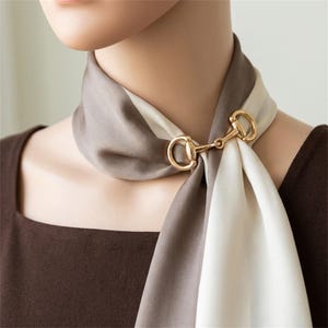 May include: A two-toned silk scarf in taupe and cream, secured at the neck with a polished gold-tone metal horsebit clasp. The smooth fabric drapes elegantly around the neck, featuring a clean, solid color block design with a metallic accent.
