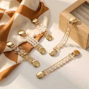 Könnte beinhalten: Vier goldfarbene Metall-Cardigan-Clips mit verschiedenen Designs: einer mit floralem Filigranmuster, zwei mit Reihen aus weißen Kunstperlen und einer mit zwei zarten Goldketten, die mit kleinen weißen Kunstperlen besetzt sind, alle mit sicheren Verschlüssen.