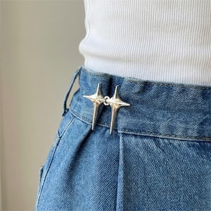 May include: A pair of silver-toned metal star-shaped fasteners attached to the waistband of a blue denim skirt. The fasteners are positioned at the centre front, connecting the two sides of the waistband with a polished, reflective finish.