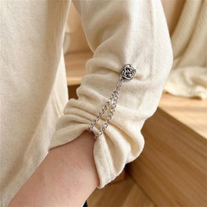May include: A cream-coloured long-sleeved knit top featuring a silver-toned metal embellishment on the sleeve. The decorative piece consists of a circular floral-shaped button connected to two delicate silver chains that drape over the fabric.