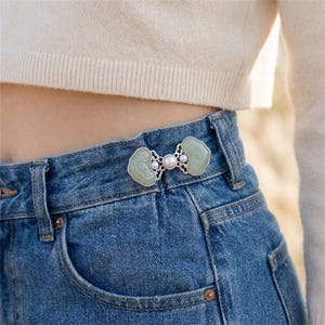 May include: A silver-toned metal waist tightener pin featuring two pale green, cloud-shaped jade-like stones and three small white pearls. The decorative fastener is attached to the waistband of blue denim jeans, cinching the fabric together.