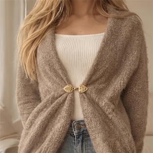 Könnte beinhalten: Eine beigefarbene, flauschige Strickjacke mit offener Vorderseite und einem goldfarbenen Metallverschluss, der aus zwei ineinandergreifenden herzförmigen Elementen besteht. Die Jacke wird über einem weißen Ripp-Top und einer blauen Jeans getragen.