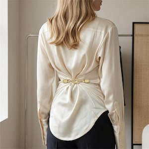 Puede incluir: Una blusa de satén color crema de manga larga con un clip de metal dorado en la parte central de la espalda. El clip tiene un diseño de nudo decorativo y frunce la tela para crear una silueta entallada. La blusa tiene puños abotonados.