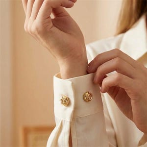 May include: A cream-colored long-sleeve shirt featuring a structured cuff with two gold-toned buttons. Each button is shaped like a small, detailed rose. The fabric of the shirt appears smooth and lightweight, with visible pleats near the cuff area.