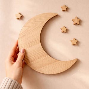 May include: Wooden crescent moon and star wall decor. The moon is a light brown color and is surrounded by five wooden stars. The decor is designed to be mounted on a wall. The background is a light beige color.