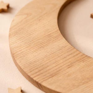 May include: Close-up of a wooden crescent moon with visible wood grain. The light brown moon shape is surrounded by a soft, neutral background. Small wooden star shapes are scattered around the moon.