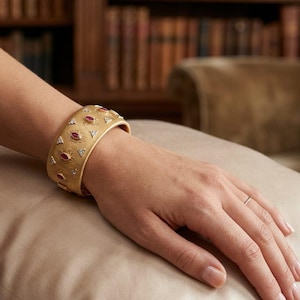 Könnte beinhalten: Ein goldfarbenes Armreif-Armband mit ovalen roten Edelsteinen und kleinen Diamantakzenten. Das Armband wird am Handgelenk getragen und liegt auf einer beigen Oberfläche. Der Hintergrund umfasst ein Bücherregal und einen Stuhl.