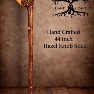 Pode incluir: Um bastão de avelã de 44 polegadas com um topo arredondado de cor clara e um eixo texturizado coberto por casca castanha escura. O texto "HAND CRAFTED" aparece acima de um gráfico de árvore, seguido de "Hand Crafted 44 inch Hazel Knob Stick."