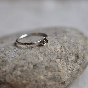 May include: A delicate silver ring with a small, flower-like design. The ring band has a hammered texture, and the focal point is a tiny, rounded bloom. The ring is displayed on a grey stone, showcasing its minimalist style.