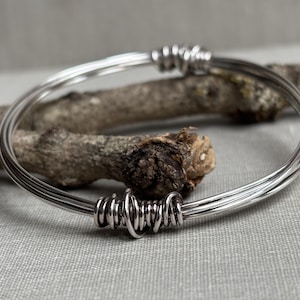 Op de afbeelding: Een zilverkleurige armband met een multi-strand ontwerp. De armband is met tussenpozen met draad omwikkeld. De achtergrond is voorzien van een stuk hout op een grijze ondergrond.