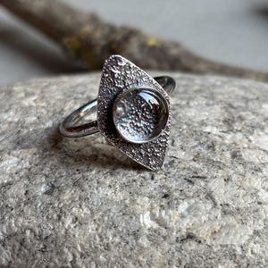 Puede incluir: Un anillo de plata con un engaste texturizado en forma de diamante. El engaste contiene una piedra redonda y transparente. El anillo se muestra sobre una superficie de piedra gris, mostrando su diseño único y artesanía.