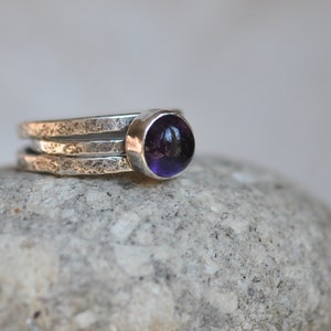Puede incluir: Un anillo de plata con tres bandas, una de las cuales sostiene una piedra preciosa redonda de color púrpura intenso. El anillo tiene un aspecto texturizado y martillado. La piedra preciosa es de un color púrpura intenso y rico.