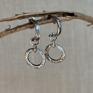 May include: A pair of silver hoop earrings with a textured finish. Each earring features a smaller, textured circle dangling from the hoop. The earrings are displayed against a neutral background, highlighting their metallic sheen and intricate design.