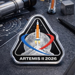 May include: A triangular patch featuring a rocket launching from the moon, with blue and red swooshes. The patch has the text "ARTEMIS II 2026" at the bottom. The patch is black with a silver border.