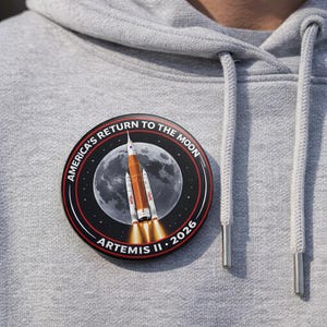 May include: A round, black and red Artemis II 2026 patch with a rocket launching towards the moon. The patch reads "AMERICA'S RETURN TO THE MOON" in white text. The patch is on a gray hoodie.