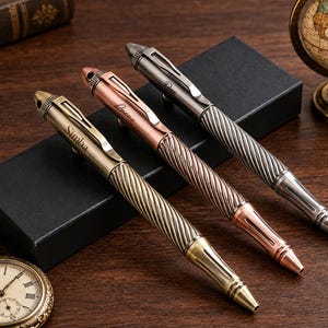 以下が含まれることがあります： ツイストバレルデザインとキャップにクリップが付いた3本の金属製ペン。アンティーク真鍮、銅、ガンメタルグレーの仕上げ。各ペンにはキャップに名前が刻印されており、Simba、Lucna、および3番目の名前が確認できます。
