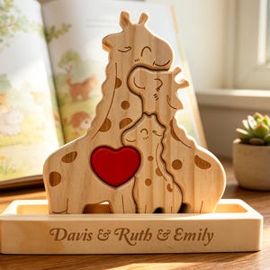 Op de afbeelding: Een lichtgekleurde houten puzzel met drie in elkaar grijpende giraffefiguren en een felrode hartvormige uitsparing in het midden. De puzzel rust op een rechthoekige houten voet met de namen Davis & Ruth & Emily in sierletters.