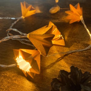 Könnte beinhalten: Eine Lichterkette mit gefalteten, sternförmigen Papierschirmen in einem warmen Bernsteinton. Die Schirme werden von innen beleuchtet und werfen einen sanften Schein auf das dünne, transparente Stromkabel. Eine kleine Eichel und ein Tannenzapfen liegen daneben.