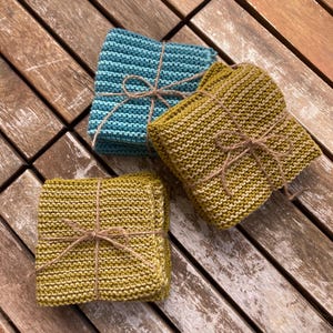 Könnte beinhalten: Drei gestrickte Waschlappen in Türkis und Olivgrün, mit Bindfaden zusammengebunden. Die Waschlappen sind gestapelt und scheinen handgefertigt zu sein. Der Hintergrund ist eine Holzoberfläche.