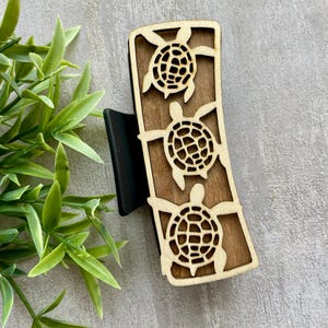 May include: Wooden hair clip featuring three laser-cut sea turtles. The turtles are light brown against a darker brown background. The clip has a black base and a curved rectangular shape. The design is suitable for various hair types and styles.