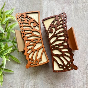 Peut inclure: Deux pinces à cheveux rectangulaires avec un revêtement en bois brun foncé découpé au laser représentant un motif d'aile de papillon. Elles ont une base crème et des composants latéraux en plastique beige pour le mécanisme.