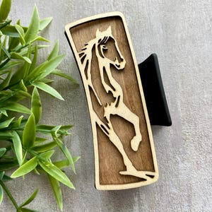 May include: A rectangular wooden hair clip featuring a cutout design of a horse. The horse is depicted in a rearing pose, with intricate details. The clip has a dark brown border and a black clip mechanism.