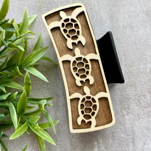 May include: Wooden hair clip featuring three laser-cut sea turtles. The clip has a dark brown wooden base with light brown turtle cutouts and a black clip mechanism. The design is set against a neutral background with green foliage.