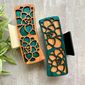 May include: Two wooden hair claw clips with a laser-cut succulent design. One clip has a natural wood finish with teal backing, the other is teal with a natural wood finish. The clips have a rectangular shape with a curved top.