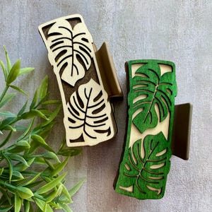 Peut inclure: Deux pinces à cheveux en bois avec un motif de feuilles de monstera. L'une est de couleur bois naturel, l'autre est peinte en vert. Les pinces ont une forme rectangulaire avec un motif de feuilles découpées et un fermoir marron.