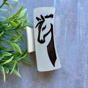 May include: A wooden hair clip with a horse head design. The horse's head is depicted in a dark brown color against a light wood background. The clip has a curved shape and a smaller, off-white piece attached to the back.